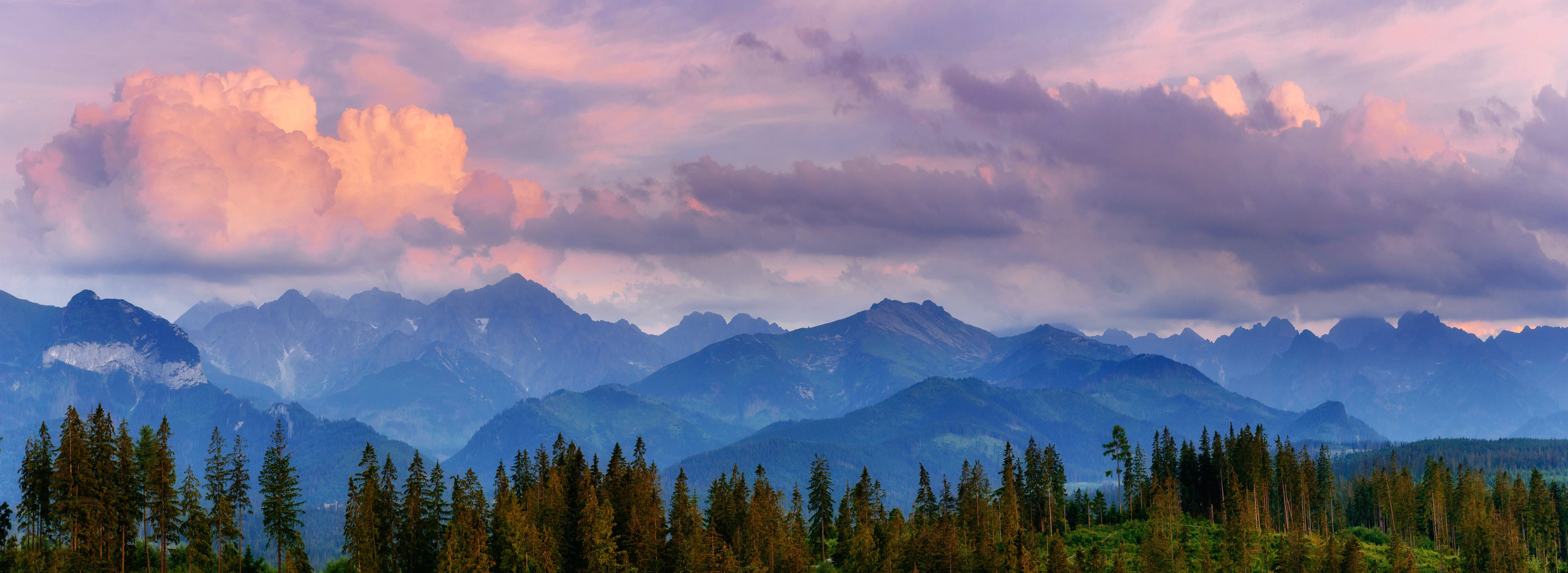 View of snowy mountains with trees in foreground and sunset cloudy skies in the background